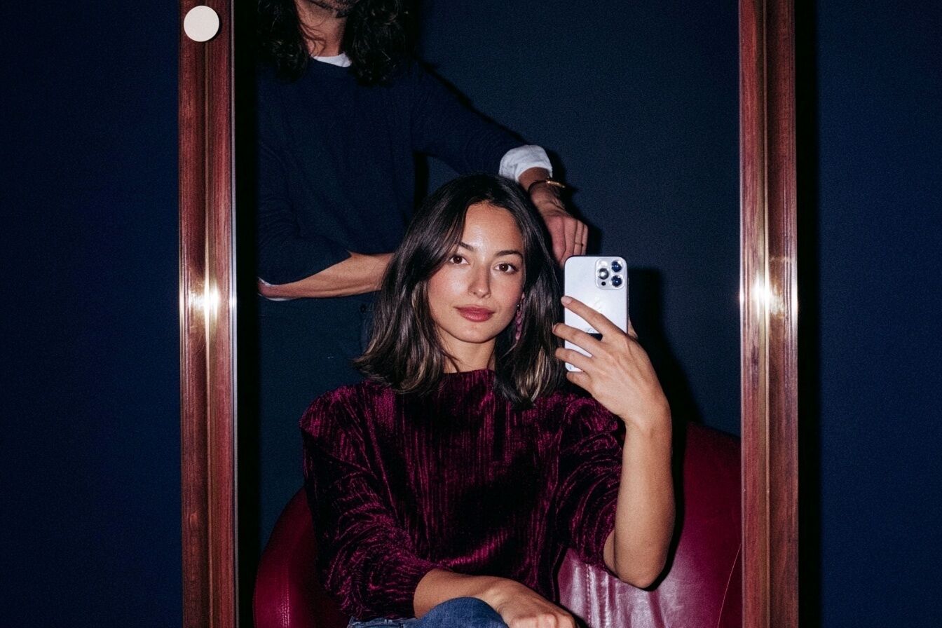 Eine Kundin macht nach dem Friseurbesuch ein Spiegel-Selfie, während der Stylist ihren Look vollendet.